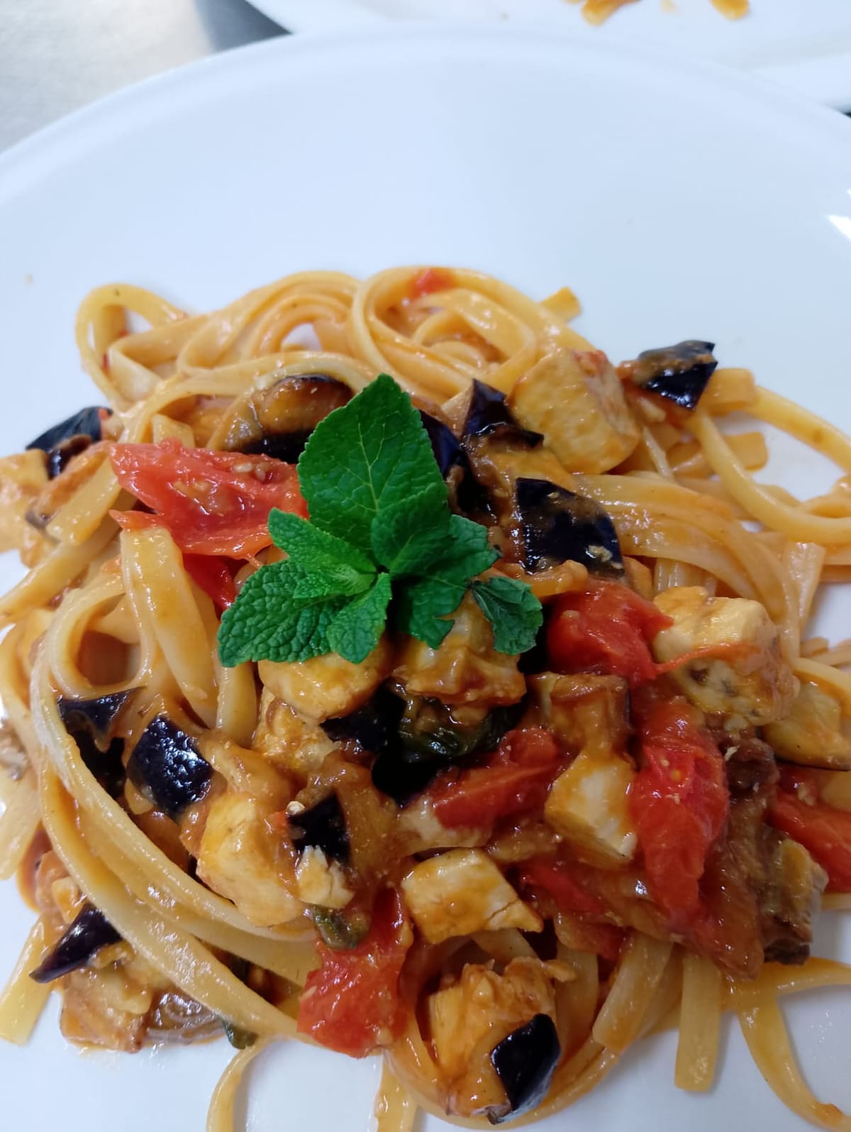 Piatto di linguine con melanzane a cubetti, pomodoro fresco e una foglia di menta sopra.