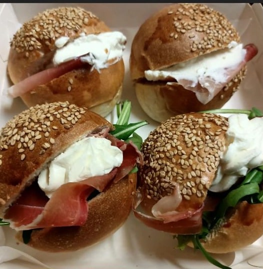 Quattro paninetti al sesamo aperti, riempiti con prosciutto crudo, formaggio fresco e rucola, nella vaschetta.