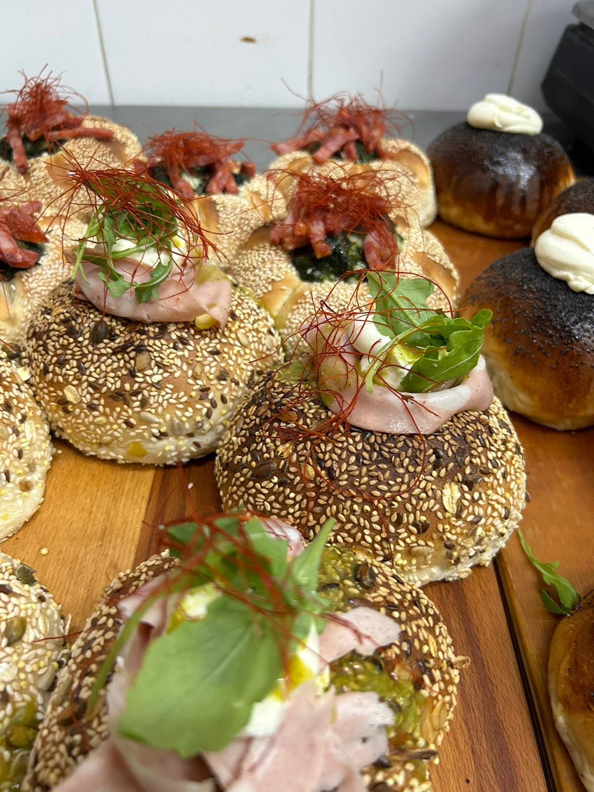 Panini ai semi di sesamo, papavero e lino, farciti con mortadella, formaggio fresco, pesto, rucola e filetti di peperoncino, su un tagliere di legno.