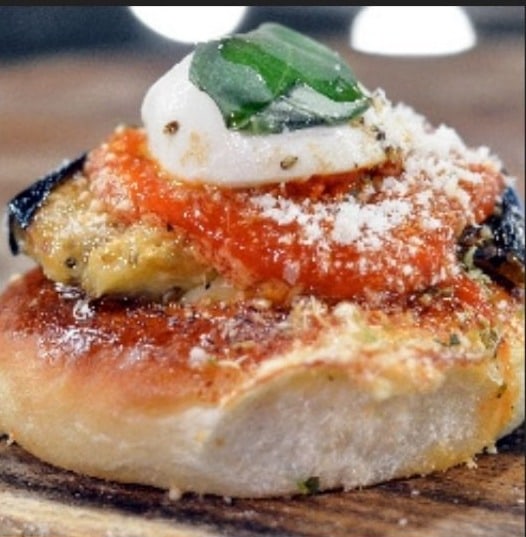 Pizzetta con una fetta di melanzana grigliata, pomodoro, formaggio fresco e una foglia di basilico sopra.