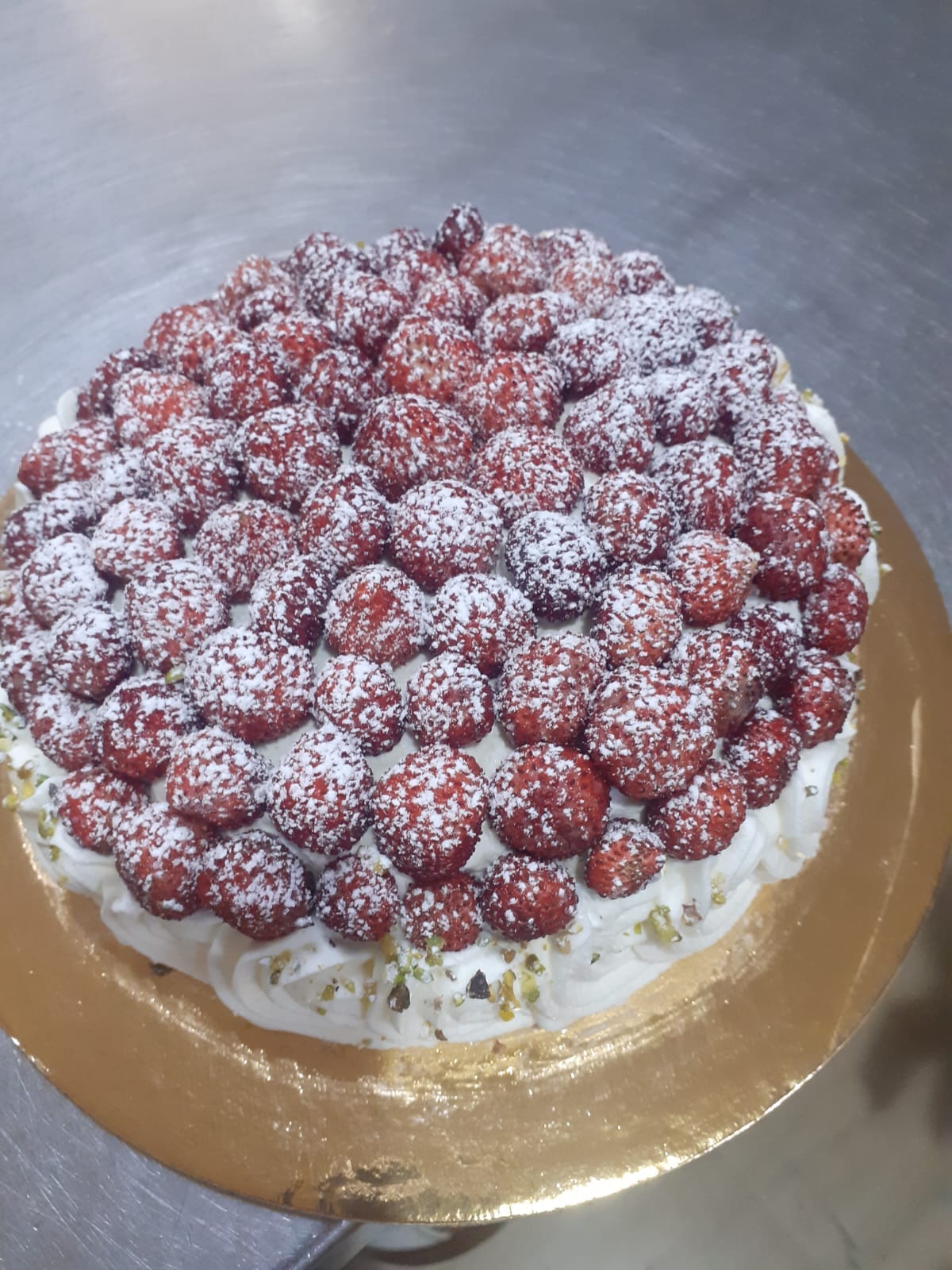Torta rotonda coperta di fragoline fresche spolverate di zucchero a velo, con il bordo di panna montata a ciuffi e la granella di pistacchio, sul vassoio dorato.