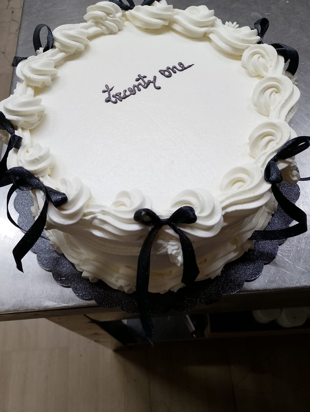 Torta rotonda con la panna montata a ciuffi tutto intorno, la scritta twenty one in cioccolato e i fiocchi neri sul bordo.