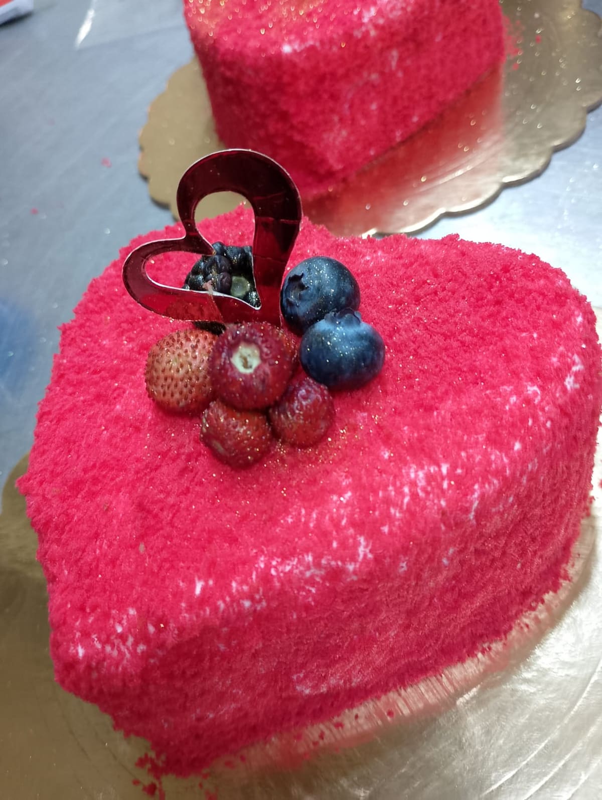 Torta a cuore ricoperta di velluto rosa acceso, con fragole, mirtilli e una decorazione a cuore sopra, sul vassoio dorato.
